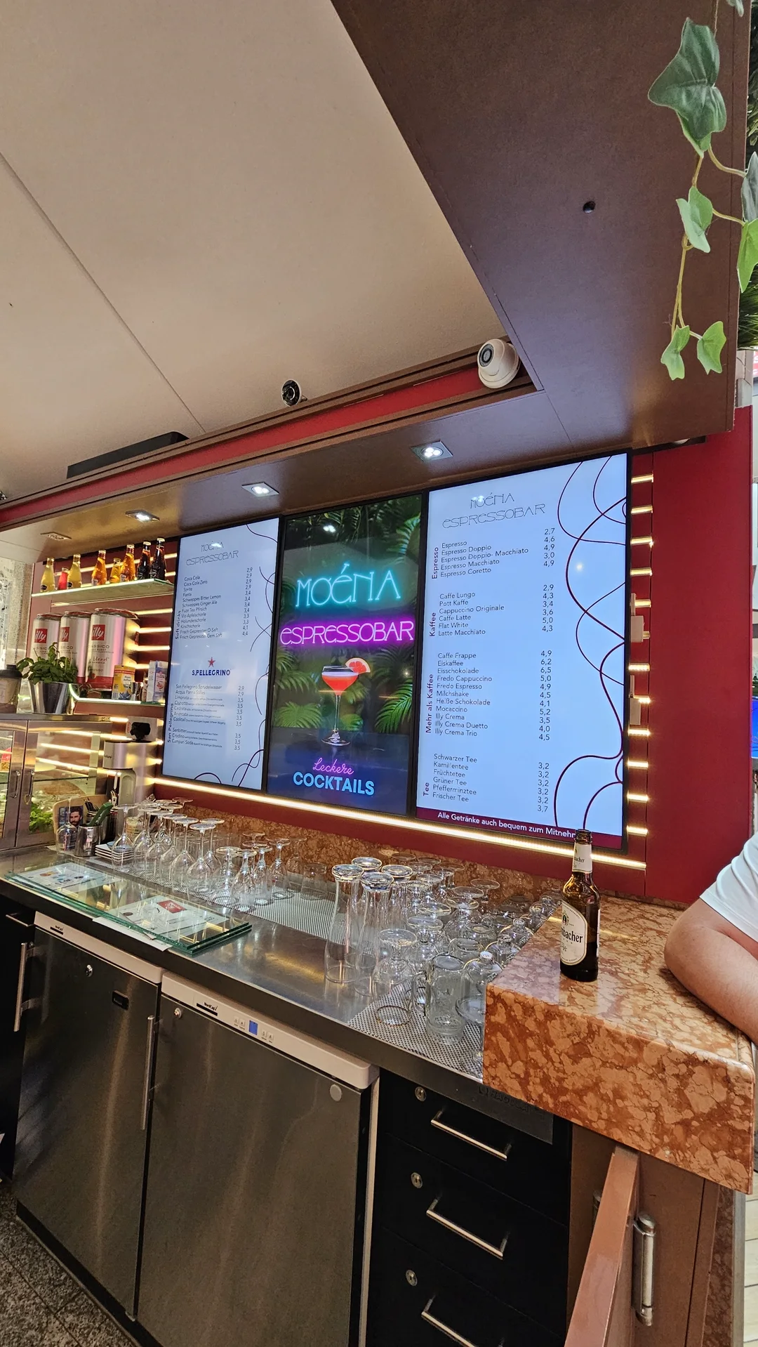 Bar der Moena Espressobar mit drei digitalen Stelen, in der Mitte das Neon-Motiv „Moena Espressobar – leckere Cocktails“