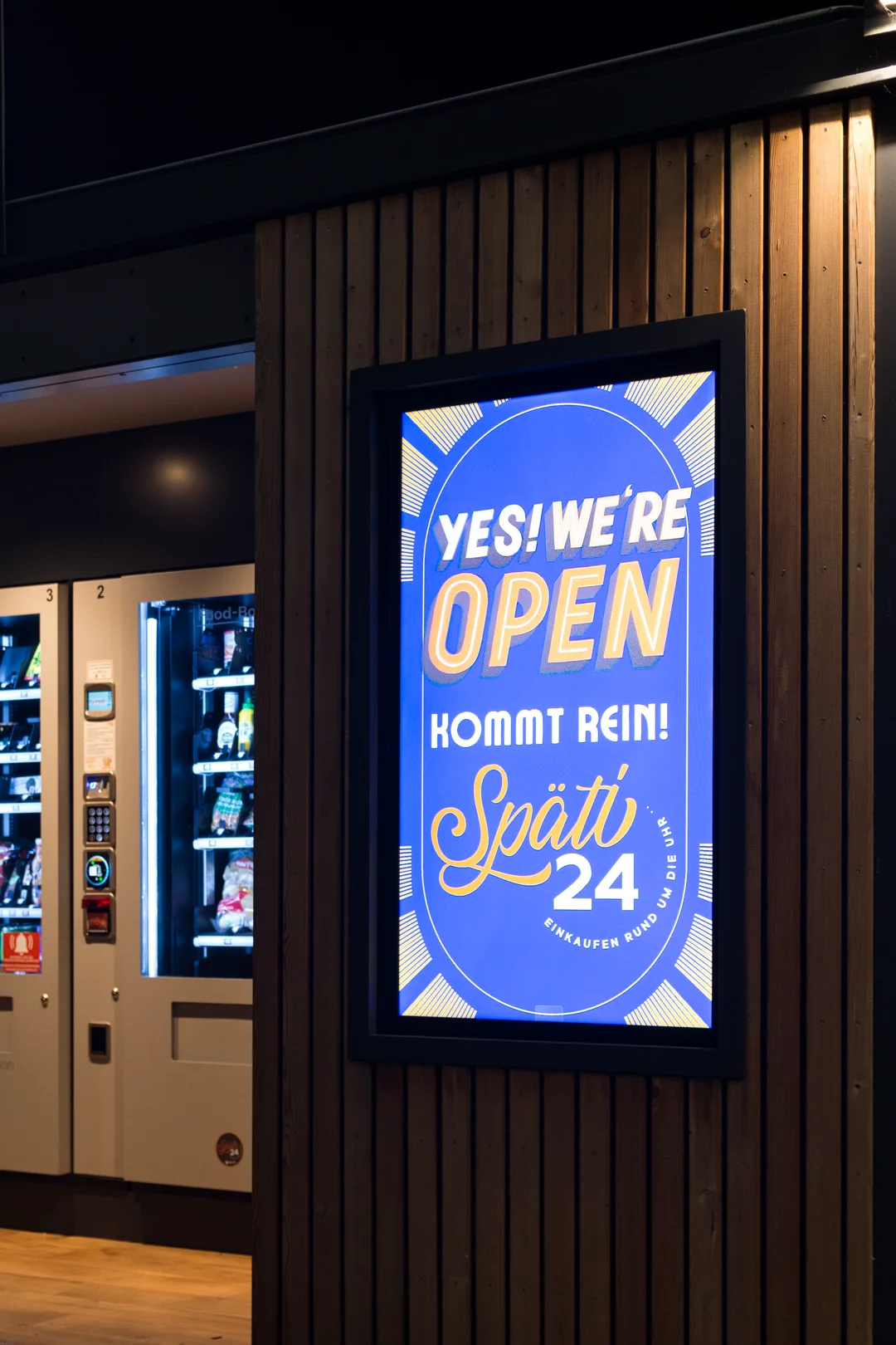 Außendisplay von Späti 24 bei Nacht mit dem Motiv „Yes! We're open – kommt rein!“, daneben die Verkaufsautomaten